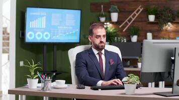 Businessman early at work alone in the office types on computer. Modern design with wooden wall and green plants. Big TV and monitors displaying charts and statistics video