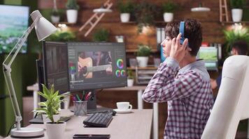 Female video editor and colorist puts her headphones on. In the background - professional post production house with a team working