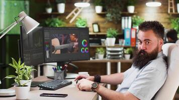 Portrait of male video editor or colorist working in cozy and stylish office. He has a dual screen setup and uses software modern software