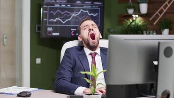 Businessman yawning while typing on the computer in modern office with big TV displaying sales data video