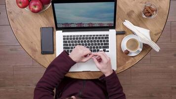 Top view of a man drinking coffee at a wooden table. Man in a coffee shop watching videos at his laptop.