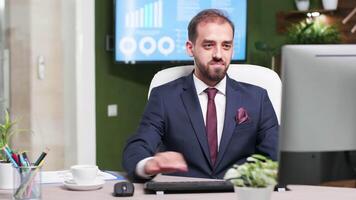 Smiling sales person in business suit working on computer in modern office with creative design. TV in the background displays sales statistics video