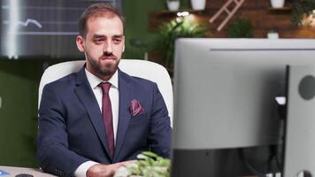 Confident office worker typing on the computer and smiling. He is alone in modern office with creative design. In the background - TV and monitors displaying graphs and sales video