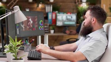 Cool looking bearded videographer or colorist works on a project in professional software. In the background - his colleague is working on other footage video