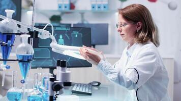 Chemist woman analysis a sample using a big magnifying glass. In the background - modern research facility with an active team video