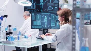 In modern research facility scientist woman works on the computer at her desk. In the background - her team is working on computers and tubes with liquid video
