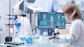 Female researcher typing on computer keyboard and looking in a microscope. In the background - chemist team actively works video