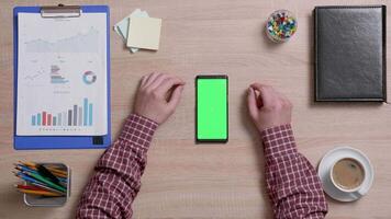 Top view of male hands scrolling on the top edge of a smart phone with green screen on. Above shot of work environment concept. Chroma key. Business and finance. video