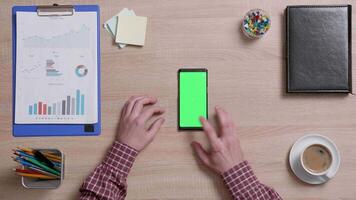 Top view of a man's hands touch the bottom left corner of a green screen on a smart phone. Above shot of work environment concept. Chroma key. Business and finance. video