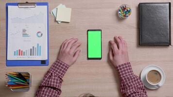 Top view of male hands and a smart phone with green screen on a wooden office desk. Above shot of work environment concept. Chroma key. Business and finance. video