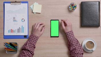 Top view of male hands scrolling on the right edge of a smart phone with green screen on. Above shot of work environment concept. Chroma key. Business and finance. video