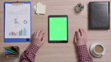 Top view of a man tap multiple times on the left bottom corner of a green screen on tablet. Office work concept. Above shot of a wooden desk. video