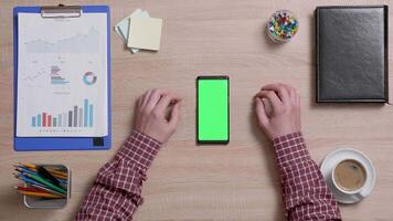 Top view of male hands scrolling on the left edge of a smart phone with green screen on. Above shot of work environment concept. Chroma key. Business and finance. video