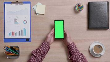 Top view of a man's hands holding his smartphone and looking at a green screen. Above shot of work environment concept. Chroma key. Business and finance. video