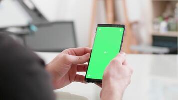 uomo si siede a un' scrivania detiene il suo smartphone con verde schermo Su. guardare a il Telefono schermo mentre a opera. ufficio concetto. video