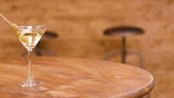 A glass of martini with olives on a wooden table. Alcoholic beverage on an empty table in front of a bar. video