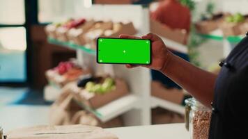 African american owner using greenscreen display at checkout, holding isolated mockup template while trying to sell organic produce at local shop. Merchant showing chromakey layout. video