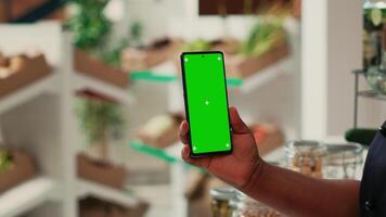 Vendor showing phone with greenscreen layout at supermarket, holding blank copyspace mockup on display. African american seller working at local grocery store, isolated chromakey screen. video