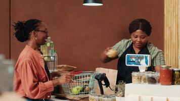 africain américain client approchant check-out avec divers marchandises, achat Frais mûr des fruits et des légumes. souriant femme demander marchande à propos éthiquement source biologique des produits sur afficher. video