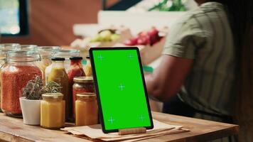 Tablet with greenscreen display placed on farmers market counter, next to homemade additives free sauces and cooking products in recyclable jars. Merchant displaying blank mockup template. video