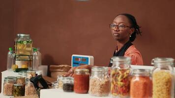 Vendor using electronic scale at checkout counter, selling organic homemade produce for vegan client at local supermarket. African american merchant putting natural fresh merchandise in a paper bag. video