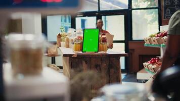 comerciante muestra bio productos siguiente a pantalla verde tableta, trabajando a un local tienda de comestibles tienda y agricultura. cliente comprobación pasta en frascos, artilugio corriendo copyspace aislado monitor disposición. trípode disparo. video