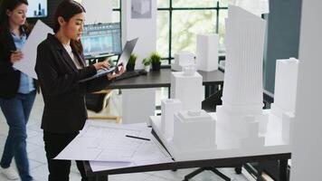 Asian contractor takes notes about 3d printed model on laptop, examining outline and scale of building maquette in architectural agency office. Cad specialist measuring layout for new project. video