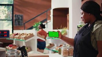 Storekeeper holds mobile phone with greenscreen display while she stands at the checkout counter. African american merchant showing isolated chromakey layout on mobile phone app. Tripod shot. video