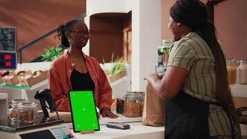 Woman approaching checkout with greenscreen on tablet, buying natural food from local vendor. African american seller using isolated mockup layout on gadget, chatting with customer. Tripod shot. video