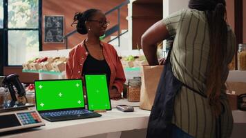 Client buying bio produce next to greenscreen at cash register, supporting farming small business. Storekeeper using blank copyspace display on laptop and tablet placed on counter. Tripod shot. video