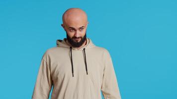 Middle eastern man acting casual on camera and showing groovy dance moves, feeling relaxed and carefree with beige hoodie. Young adult dancer having fun over blue background in studio. Handheld shot. video