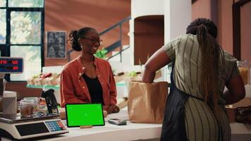 Customer approaching counter with greenscreen on device, buying eco friendly goods from local owner. African american vendor using isolated copyspace display on tablet, talks to client. Tripod shot. video