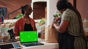 Retailer having laptop that shows greenscreen on display at cash register, giving eco products to client at market. Woman using computer with isolated chromakey template, mockup layout. Tripod shot. video