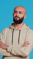 Vertical Video Middle eastern confident person posing on studio camera, feeling serious and neutral over blue background. Arab model being relaxed with beige hoodie, smiling on backdrop. Camera 2.