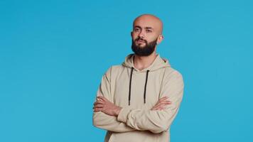 Middle eastern confident person posing on studio camera, feeling serious and neutral over blue background. Arab model being relaxed with beige hoodie, smiling on backdrop. Camera 2. video
