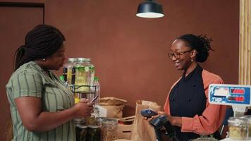 Vegan buyer paying with mobile phone at pos terminal, purchasing organic produce and additives free bulk items. African american client using nfc payment to buy fruits and vegetables. Handheld shot. video