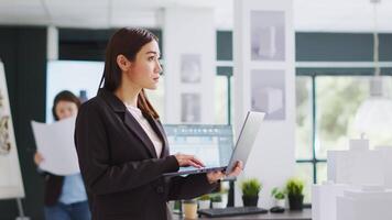 sviluppatore fa schizzi su il computer portatile per costruzione programma mentre ispezionando 3d edificio modello su design tavolo nel coworking spazio. contraente sviluppando rinnovamento schema e generando planimetria. video