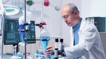 In modern and high end laboratory, doctor in his 60s is looking at different tubes with blue samples. His team is walking in the background video