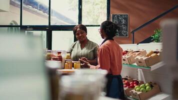 Retailer showing natural homemade sauces in reusable jars, recommending organic products to support vegan nutrition. Buyer looking at various bulk items at local farmers market. Handheld shot. video