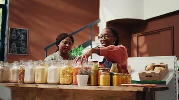 Vendor presents bio homemade sauces in reusable jars, recommending organic pantry supplies to support healthy nutrition. Client looking at various bulk items. Handheld shot. video