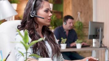 Close up portrait of female sales representative talking with a client through a headset. Busy but cozy office video