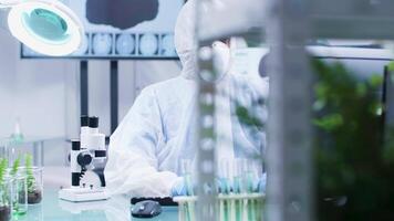 Biologist dressed in protective coverall and wearing a mask is lookign at a microscope while working in modern research facility video