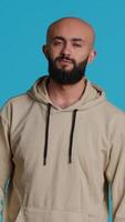 Vertical Video Middle eastern confident person posing on studio camera, feeling serious and neutral over blue background. Arab model being relaxed with beige hoodie, smiling on backdrop. Camera 1.