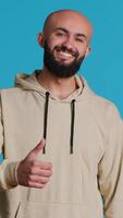 Vertical Video Middle eastern person does thumbs up sign on camera, expressing his agreement over blue background. Arab model showing satisfaction and okay symbol, positive gesture. Camera 1.