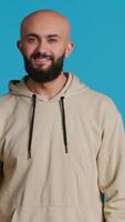 Vertical Video Middle eastern guy greeting people in front of camera, waving hello and smiling over blue background. Young person posing with confidence in studio, feeling confident and joyful. Camera 1.