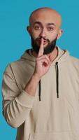 Vertical Video Middle eastern man doing hush silence symbol in studio, asking to keep a secret over blue background. Arab person creating mute gesture to be quiet in front of camera. Camera 1.