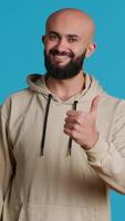 Vertical Video Arab male model gives a thumbs up in studio, expressing his satisfaction over blue background. Middle eastern guy showing approval and like gesture, positive symbol. Camera 1.