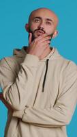 Vertical Video Thoughtful person being puzzled about a decision, standing over blue background in studio and thinking. Pensive middle eastern man being uncertain and doubtful about something. Camera 1.