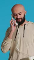 Vertical Video Muslim adult taking call on a landline phone in studio, using vintage office telephone with a cord. Arab guy standing over blue background and having a discussion on a remote call. Camera 1.