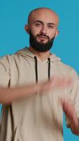 Vertical Video Middle eastern person doing timeout gesture on camera, asking for a break to stop working over blue background. Muslim adult presents symbol to pause or stop something. Camera 1.